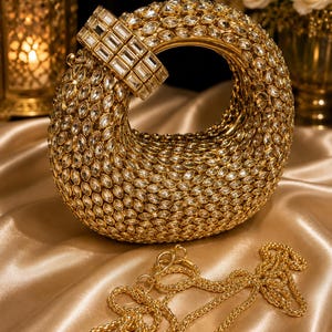 May include: A gold-toned, crescent-shaped clutch bag fully encrusted with clear, marquise-cut crystals. The bag features a rigid, circular handle adorned with rectangular crystals and includes a detachable gold-toned metal chain strap.