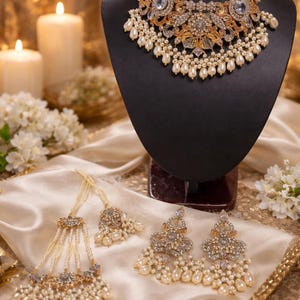 May include: An ornate necklace, earrings, and a headpiece set on a black bust. The jewelry features gold-toned metal, clear crystals, and faux pearls. The necklace is the focal point, with a large pendant. The set is displayed on a satin cloth.