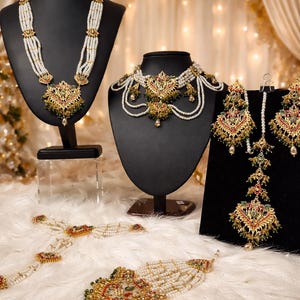 May include: An array of ornate, gold-toned necklaces, earrings, and a bracelet, adorned with pearls and colorful gemstones. The jewelry is displayed on black mannequins and a white, fluffy surface, with a soft, warm background.