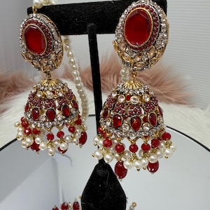 May include: Ornate gold-coloured earrings with large, oval, red gemstones. The earrings have a bell-shaped design, decorated with red and clear gemstones, and dangling white pearls. A matching necklace is also visible.