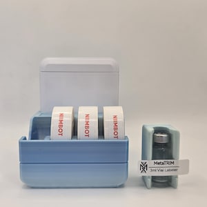 May include: A blue and white 3D-printed organiser with three rolls of white tape labelled "NIIMBOT" in red. A small vial is held in a separate blue holder, with a label that reads "MetaTRIM 3ml Vial Labeler."