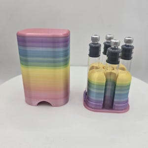 May include: A 3D-printed storage case with a rainbow-colored gradient finish, featuring a removable lid and a base that holds four small, clear glass vials with silver-colored metal caps and black rubber stoppers, arranged in a square formation.