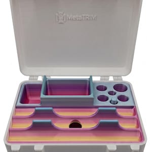 May include: A white plastic storage case with yellow latches, featuring the embossed text MetaTRIM on the interior lid. The interior contains a multi-compartment organizer insert with a gradient color pattern of pink, yellow, and blue.