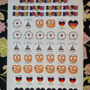 May include: A sheet of stickers featuring German-themed designs. The stickers include phrases like "BRAVO! SUPER!" and "TOLL!" alongside images of pretzels, hearts in German flag colours, and castles. The stickers are round and rectangular.