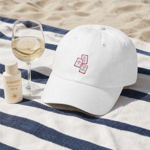 May include: A white baseball cap featuring an embroidered design of three mahjong tiles on the front. The tiles are depicted with red, green, and white colors, showcasing specific characters and symbols. The cap has a structured crown and a curved brim.
