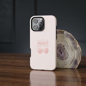 May include: A light pink phone case featuring a cherry design with a bow. The case is shown on a wooden surface next to a decorative stone. The phone case is designed to fit a smartphone.