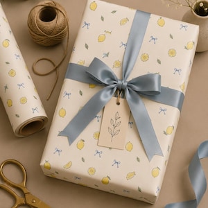 Pode incluir: Uma caixa de presente retangular embrulhada em papel creme com um padrão de limões amarelos, folhas verdes e pequenos laços azuis. A caixa está atada com uma fita de cetim larga azul ardósia e tem uma etiqueta com um desenho de uma folha.