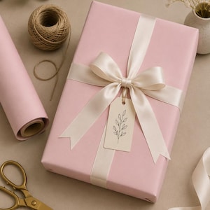 May include: A wrapped gift in pink paper with a cream-colored ribbon and bow. A small tag with a botanical illustration hangs from the bow. Gold scissors, a roll of pink paper, and twine are also visible.