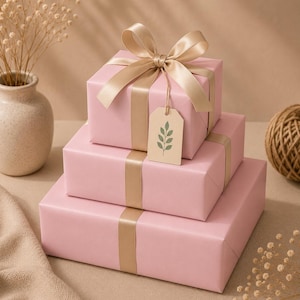 May include: Three stacked pink gift boxes with gold ribbon and bows. A small tag with a green leaf design hangs from the top box. The boxes are on a neutral surface with a vase of dried flowers and a ball of twine.