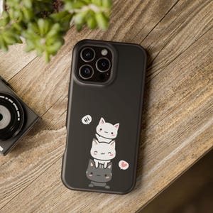 May include: Black phone case featuring a stack of three cartoon cats. The top cat says "Hi" in a speech bubble, and a heart icon is next to the bottom cat. The case is on a wooden surface.
