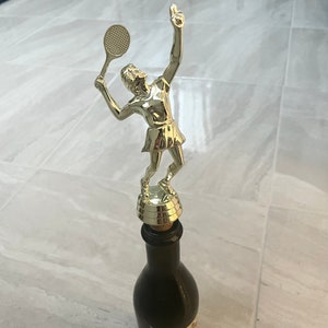 Könnte beinhalten: Eine goldfarbene Trophäe einer Tennisspielerin, die einen Schläger hält, montiert auf einer dunklen Glasflasche.