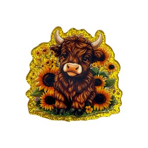 May include: A decorative pin featuring a brown Highland cow with curved horns, surrounded by sunflowers. The cow has a light-colored muzzle and is set against a glittery gold background. The pin is likely for a bag, jacket, or other accessory.
