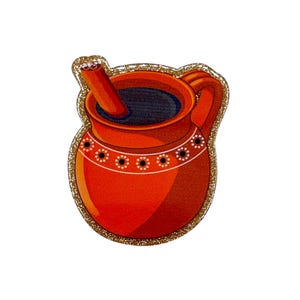 May include: A decorative, orange-red ceramic pitcher with a dark liquid inside and a wooden stirring stick. The pitcher has a handle and is outlined with gold glitter. White dots with black centers decorate the rim.