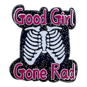 May include: A black and glittery pin with the words "Good Girl Gone Rad" in pink lettering. The pin features a white rib cage design in the center. The design is outlined in pink and glitter.