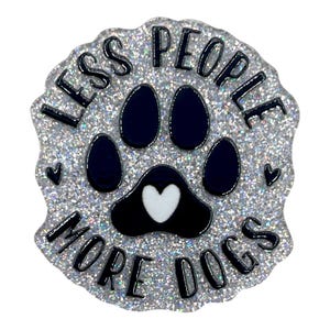 May include: A glittery, round pin with the text "LESS PEOPLE MORE DOGS" in black. The pin features a dark blue paw print with a white heart in the center. Small black hearts are also included in the design.