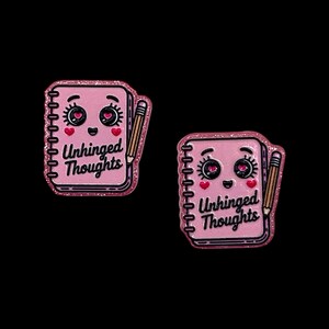 May include: Two pink enamel pins shaped like notebooks with the text "Unhinged Thoughts". Each pin has a smiling face, heart-shaped accents, and a pencil. The pins have a glittery border.
