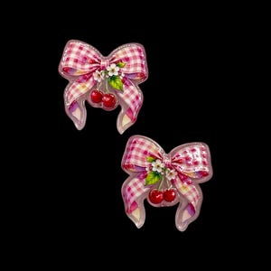 May include: Two decorative bow-shaped hair clips. Each clip features a pink and white gingham pattern, a cluster of red cherries, and small white flowers. The bows have a clear, glossy finish and are set against a black background.