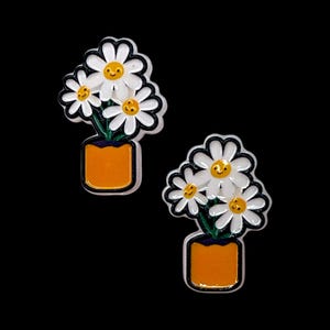 May include: Two daisy flower earrings. Each earring features a cluster of white daisies with yellow centers and smiling faces, set in an orange pot with a black outline. The background is solid black.