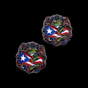 May include: Two circular metal enamel pins featuring a green frog with large eyes holding a Puerto Rican flag. The design includes intricate red, blue, and black swirling patterns surrounding the frog and flag, with a metallic silver border.