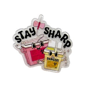 May include: A glittery, cartoon-style pin featuring a pink sharps container and a yellow biohazard container, both with smiling faces. The text "STAY SHARP" is above the containers, and a syringe is also present. The pin has a white border.