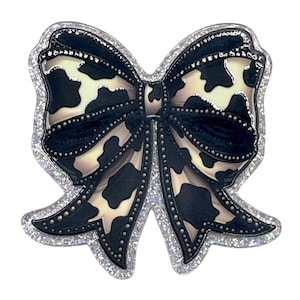 May include: A decorative bow-shaped accessory with a cow print design. The bow features a black and white pattern with a silver glitter outline and small silver studs. The bow is a fashion accessory.