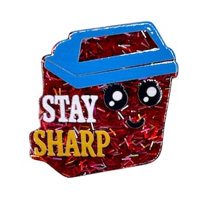 May include: A pin shaped like a smiling trash can with the words "STAY SHARP" in white and yellow. The trash can is red with glitter and a blue lid. The pin has a cute cartoon design.