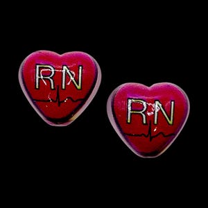 May include: A pair of heart-shaped earrings with a red background. Each earring features the letters "RN" in silver, a heartbeat line, and a dark pink border. The earrings are set against a black background.