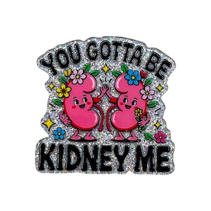 May include: A glittery enamel pin featuring two cartoon pink kidneys with smiling faces, arms, and legs. The kidneys are decorated with colorful flowers and are high-fiving. The black text reads YOU GOTTA BE KIDNEY ME in bold capital letters.