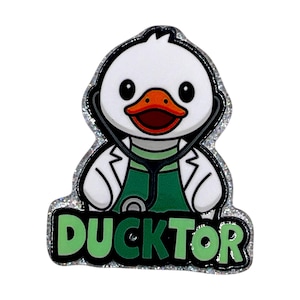 May include: A Ducktor pin featuring a cartoon duck wearing a stethoscope and a doctor's coat. The duck has a white body, an orange beak, and is outlined in black. The word "DUCKTOR" is written in green letters.
