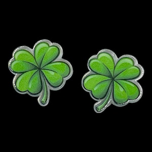 May include: Two green four-leaf clover earrings with a silver-colored border. The clovers have a gradient of green shades, with darker green veins. The background is black.