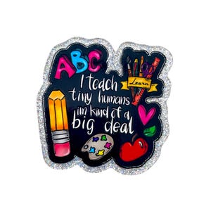 May include: A glittery, black-backed pin with the text "I teach tiny humans I'm kind of a big deal." The pin features colorful illustrations of an apple, pencil, paint palette, and scissors. The letters "ABC" are at the top.
