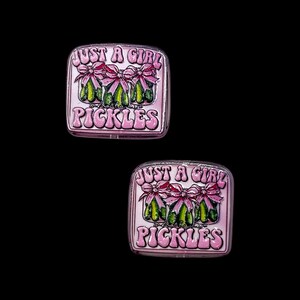 May include: Two square, pink enamel pins with the text "JUST A GIRL" above the word "PICKLES". Each pin features three pickles with pink bows. The pins have a silver border and are set against a black background.