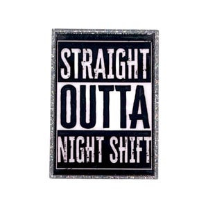 May include: A rectangular enamel pin featuring a black and white design with the text STRAIGHT OUTTA NIGHT SHIFT. The pin has a silver-toned border with a glittery finish, and the lettering is presented in a bold, distressed font style.
