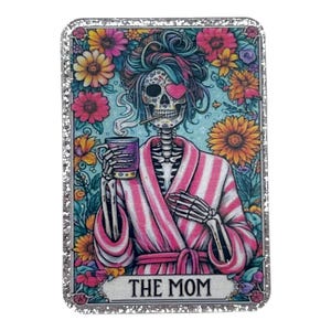 May include: A rectangular card with a glittery silver border features a skeleton in a pink and white striped robe, holding a mug and smoking. The skeleton has a heart-shaped eye patch and is surrounded by colorful flowers. The text "THE MOM" is at the bottom.