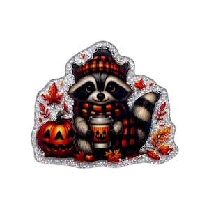 May include: A glittery sticker featuring a cartoon raccoon wearing a red and black plaid hat and scarf. The raccoon holds a cup with a jack-o'-lantern face, positioned next to a carved pumpkin and scattered autumn leaves on a silver background.