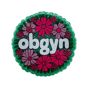 May include: A round, green sticker with a scalloped edge. The sticker features a floral design with pink and purple flowers surrounding the word "obgyn" in white, bold letters.