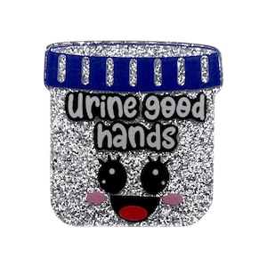 May include: A glittery, rectangular pin with a blue top and the words "Urine good hands" in black. The pin features a smiling face with black eyes, pink cheeks, and a red mouth. The background is silver.