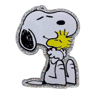 May include: A Snoopy and Woodstock character sticker. Snoopy is white with black ears and a black spot on his ear. Woodstock is yellow. The sticker has a silver glitter border and is on a white background.