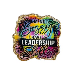 May include: A colorful pin with the text "I'M NOT Bossy I HAVE LEADERSHIP Skills". The pin has a gold glitter border and a vibrant, tie-dye-like background with leopard print accents. The text is in black and white.