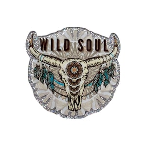 May include: A glittery sticker featuring a steer skull with long horns, decorated with teal feathers and a central mandala pattern. The text WILD SOUL is printed in brown capital letters above the skull against a beige and white background.