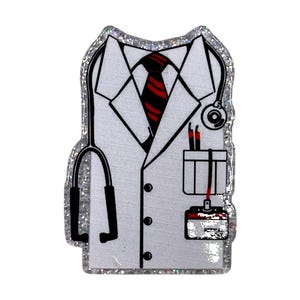 May include: A white medical coat-shaped pin with a silver glitter border. The pin features a black stethoscope, a red and black striped tie, and a pocket with pens. The design is a cartoon representation of a doctor's attire.