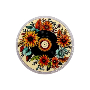 May include: A circular vinyl record graphic featuring a central black disc surrounded by vibrant orange, yellow, and red sunflowers and teal floral accents. The design is set against a cream background with a glittery, translucent outer border.