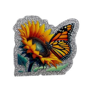 May include: A decorative pin featuring a vibrant sunflower and a monarch butterfly. The sunflower has yellow petals and a dark center, while the butterfly displays orange, black, and white patterns. The pin is outlined with silver glitter.
