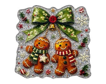 May include: A square-shaped ornament with a silver glitter background featuring two gingerbread cookies wearing striped scarves. A large green bow with holly patterns and a red button sits above the cookies, accented by small holly leaves, red berries, and white snowflakes.