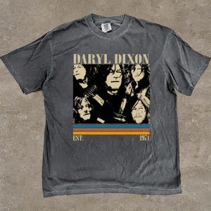 Puede incluir: Camiseta gris oscuro con un gráfico de Daryl Dixon, el texto "DARYL DIXON" encima de una serie de caras y el texto "EST. 1971" debajo. El gráfico utiliza una combinación de colores retro de naranja, azul y marrón.