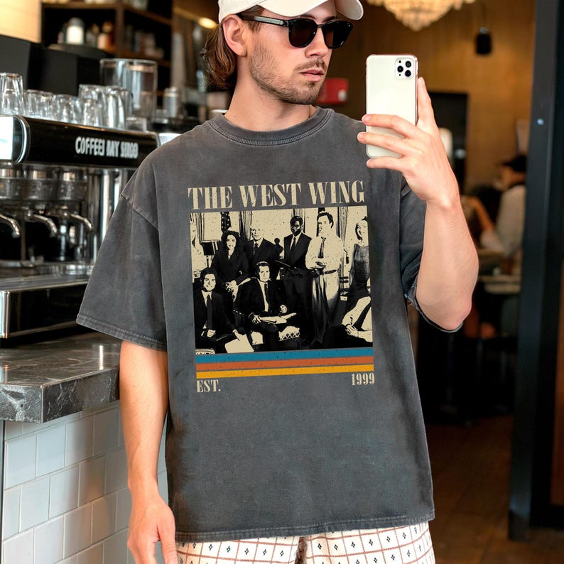 Puede incluir: Camiseta gris oscuro con un gr&aacute;fico de un grupo de personas y el texto "THE WEST WING" arriba y "EST. 1999" abajo. El gr&aacute;fico tiene un dise&ntilde;o retro con un bloque de color naranja, azul y amarillo.