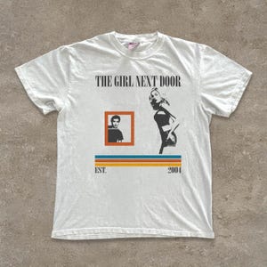 May include: White t-shirt featuring the text "THE GIRL NEXT DOOR" above a graphic of a woman and a framed image of a person. Below the images are the words "EST. 2004" above a horizontal stripe design.
