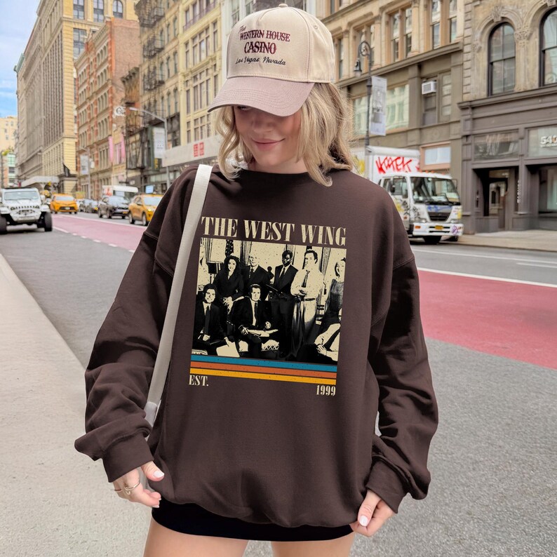 Puede incluir: Una sudadera marr&oacute;n con el texto "THE WEST WING" y una imagen en blanco y negro de un grupo de personas. Debajo de la imagen est&aacute;n las palabras "EST. 1999" y rayas horizontales. La persona tambi&eacute;n lleva una gorra de b&eacute;isbol beige.