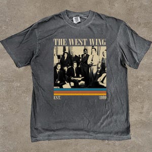 Puede incluir: Camiseta gris oscuro con un dise&ntilde;o gr&aacute;fico de la serie de televisi&oacute;n "El Ala Oeste de la Casa Blanca". El dise&ntilde;o incluye una foto grupal del elenco, el t&iacute;tulo del programa y el a&ntilde;o "1999". La camiseta tiene un aspecto vintage y desgastado.