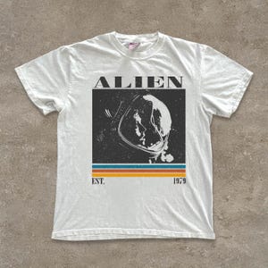 以下が含まれることがあります： 宇宙飛行士のヘルメットと「ALIEN」の文字が描かれた、かすれたグラフィックの白いTシャツ。グラフィックの下には青、オレンジ、黄色の横縞があり、その下に「EST. 1979」と書かれています。Tシャツは半袖です。